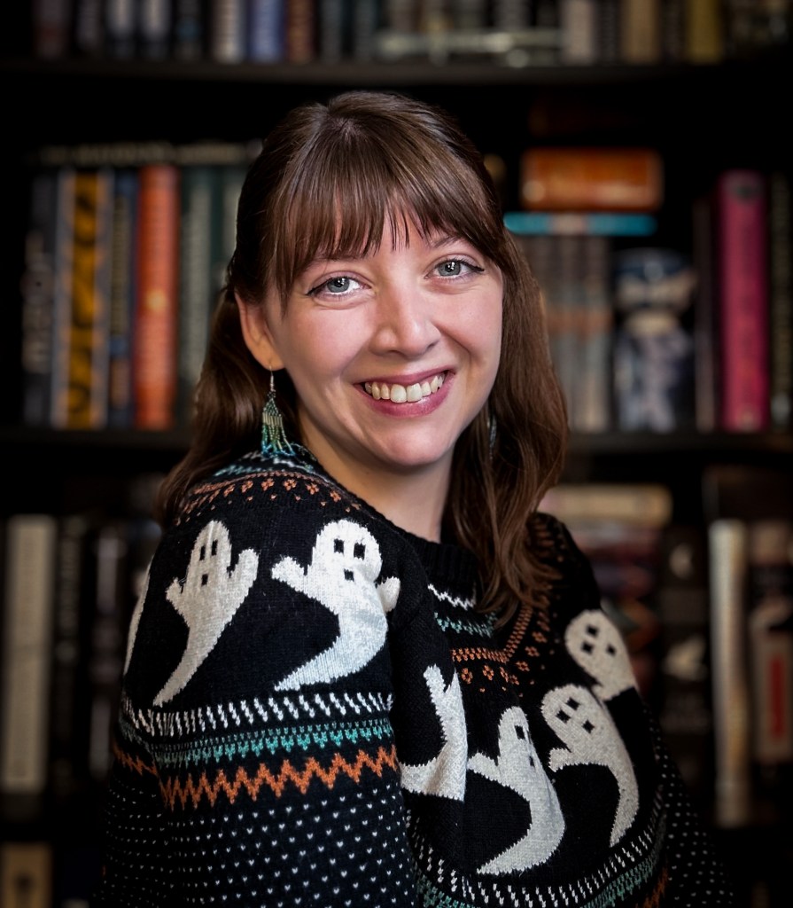 Woman with shoulder length brown hair dressed in a ghost sweater, smiling in fornt of bookshelves